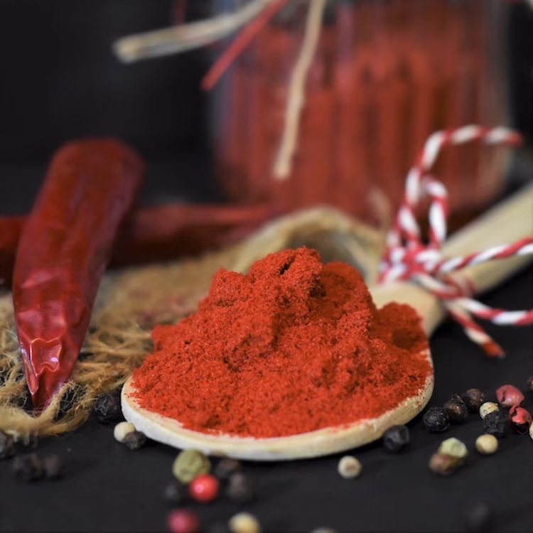 Powdered smoked paprika in a wooden spoon with red peppers and other spices scattered around.